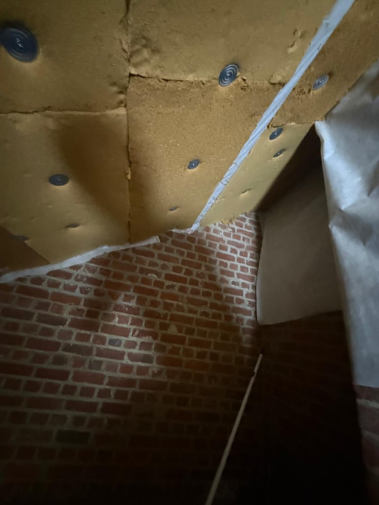 Interior construction site showing yellow insulation board on ceiling with blue anchors and white PVC pipe, above red brick flooring