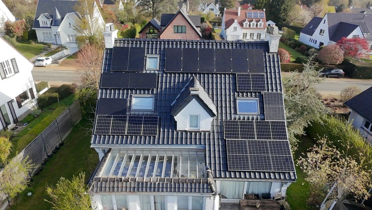 Aerial view of a residential house with extensive solar panels covering the roof, surrounded by trees and neighboring homes